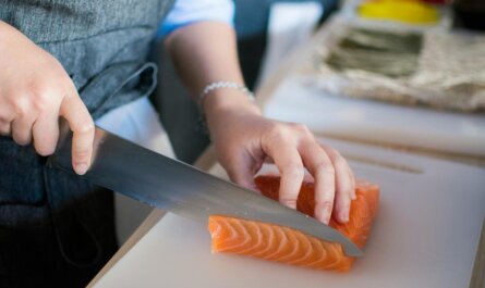 Close-up of a chef's hands slicing fresh salmon for sushi on a cutting board indoors.