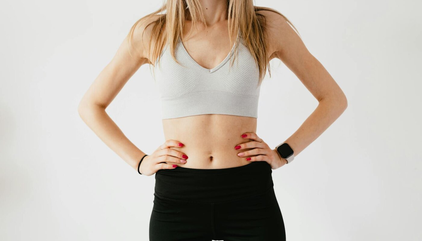 Faceless slim anonymous blond female in sports bra and black leggings in wearable bracelet showing perfect belly on white background while standing with hands on waist