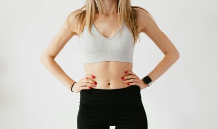 Faceless slim anonymous blond female in sports bra and black leggings in wearable bracelet showing perfect belly on white background while standing with hands on waist