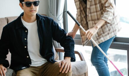 campus louis braille A visually impaired man receives support from a caregiver indoors, using a walking stick.